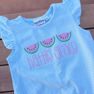 May include: Light blue and white striped romper with ruffled sleeves. The romper features embroidered watermelon slices and the name "Hattie Grace" in pink script. The tag indicates the size is 18 months.