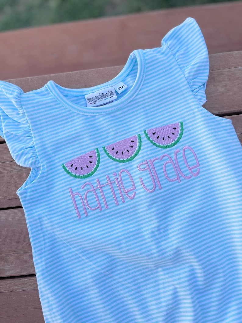 May include: Light blue and white striped romper with ruffled sleeves. The romper features embroidered watermelon slices and the name "Hattie Grace" in pink script. The tag indicates the size is 18 months.