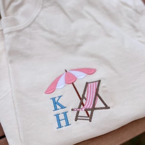 May include: A white t-shirt with a pink and white striped beach umbrella, a brown beach chair, and the letters "KH" embroidered in blue.