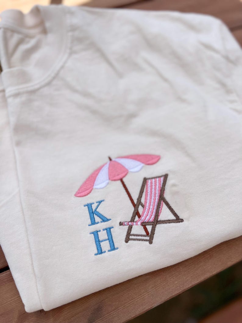 May include: A white t-shirt with a pink and white striped beach umbrella, a brown beach chair, and the letters "KH" embroidered in blue.