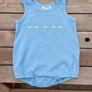 May include: Light blue baby romper with the name "KROSS" embroidered in green, with fish accents. The romper has button closures on the shoulders and snaps at the bottom. The garment is made of soft fabric.