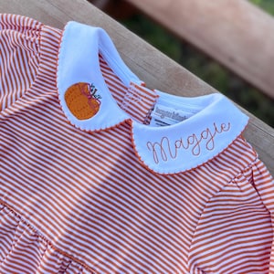 May include: A striped orange and white dress with a white collar. The collar features an embroidered orange pumpkin and the name "Maggie" in orange thread. The dress has long sleeves and a ruffled skirt, perfect for a child.