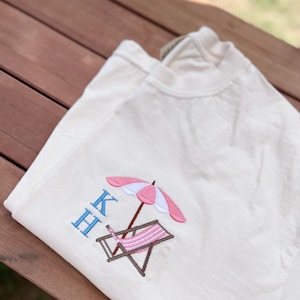 May include: A white t-shirt with a pink and white beach umbrella and a brown and white striped beach chair embroidered on the front. The letters "KH" are embroidered in blue above the beach scene.