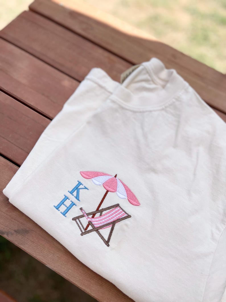 May include: A white t-shirt with a pink and white beach umbrella and a brown and white striped beach chair embroidered on the front. The letters "KH" are embroidered in blue above the beach scene.