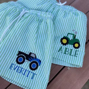 May include: Two pairs of green and white striped swim trunks with embroidered monster trucks and tractors. One pair says "Everitt" and the other says "Able".