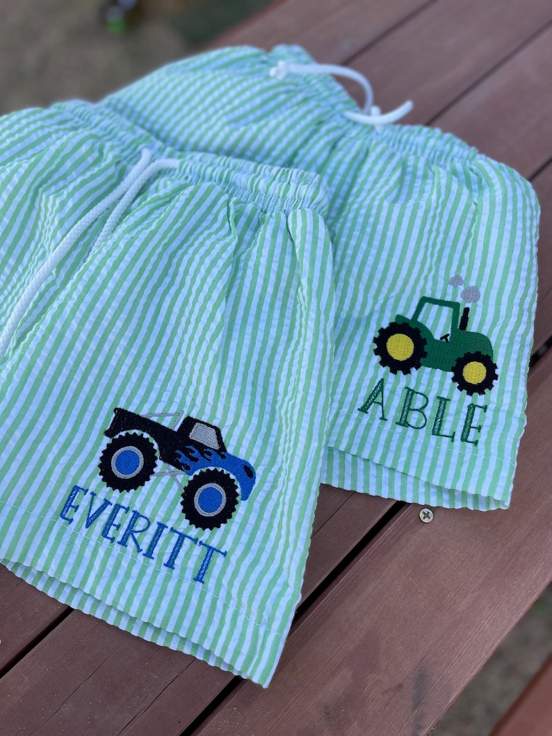 May include: Two pairs of green and white striped swim trunks with embroidered monster trucks and tractors. One pair says "Everitt" and the other says "Able".