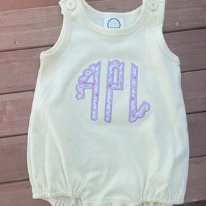 May include: A yellow sleeveless baby onesie with a floral monogram "APL" in purple and pink.