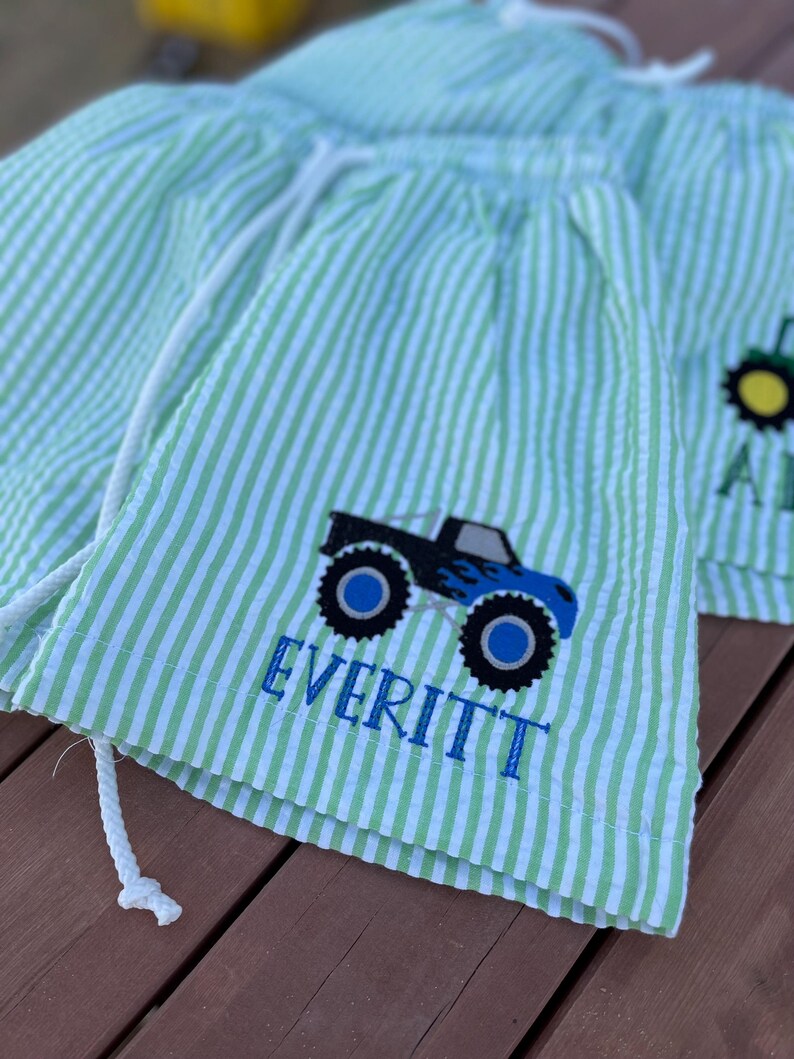 May include: Green and white striped shorts with a blue monster truck graphic and the name "EVERITT" embroidered on the front.