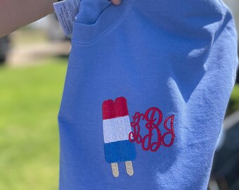 May include: A light blue sweatshirt featuring an embroidered popsicle in red, white, and blue. The sweatshirt also has a red monogram on the right side. The garment is held up by a person.