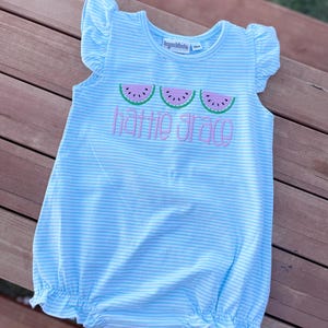 May include: A light blue and white striped baby romper with ruffled sleeves and hem. The romper features three embroidered watermelon slices and the name "Hattie Grace" in pink script. The romper is size 10m.
