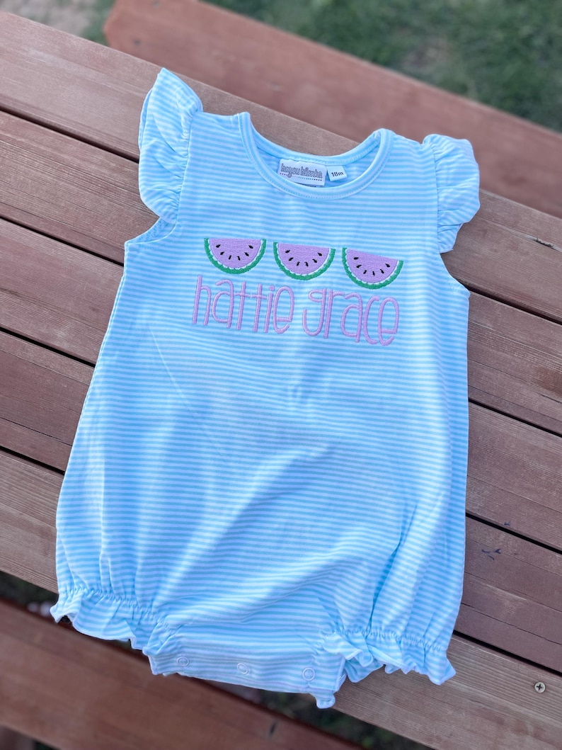 May include: A light blue and white striped baby romper with ruffled sleeves and hem. The romper features three embroidered watermelon slices and the name "Hattie Grace" in pink script. The romper is size 10m.
