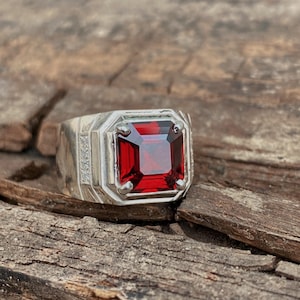 May include: A silver ring featuring a large, square, deep red gemstone. The ring has a geometric design around the stone and small accent stones on the sides. The ring is set against a rustic wooden background.