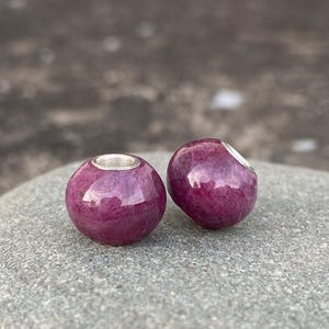 May include: Two round, polished ruby beads with silver-colored metal accents. The beads are a deep, rich magenta color with a glossy finish. The beads are likely for jewelry making.