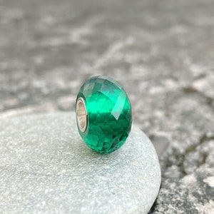 Peut inclure: Une perle de verre verte facetée avec un noyau argenté. La perle est posée sur une pierre grise.