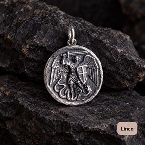Puede incluir: Un colgante circular de plata con un relieve detallado de una figura alada que sostiene una espada y un escudo mientras está de pie sobre una serpiente. El metal tiene un acabado oxidado con áreas oscuras y una anilla pulida en la parte superior.