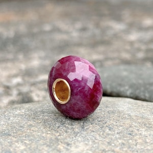 May include: A faceted, oval-shaped ruby bead with a gold-colored metal insert. The bead is a deep red color with lighter pinkish hues visible in the facets. The bead is shown on a gray stone surface.