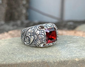 Pode incluir: Um anel de prata com uma grande pedra preciosa vermelha quadrada. O anel apresenta intrincados desenhos florais e de rolagem. A pedra preciosa é de uma cor vermelha profunda e rica, e o anel está sobre uma superfície de pedra.