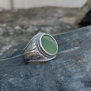 Puede incluir: Un anillo de plata con una piedra verde ovalada grande. El anillo tiene detalles grabados ornamentados en la banda, incluyendo patrones florales y de hojas. La piedra está engastada en un bisel liso.