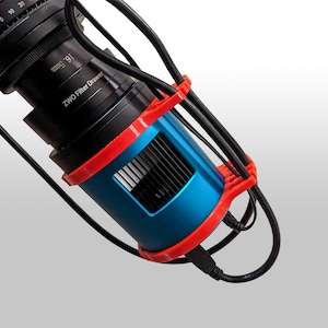 Op de afbeelding: Een zwarte en blauwe camera lens met een rode filterlade. De lens heeft een zwarte kabel eraan bevestigd en is gemonteerd op een rode en zwarte beugel.