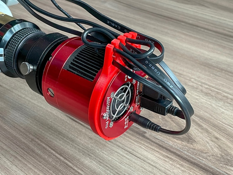 K&ouml;nnte beinhalten: Eine rote Astronomie-Kamera mit einem K&uuml;hlgebl&auml;se und mehreren angeschlossenen Kabeln. Die Kamera hat ein schwarzes Objektiv und ein Etikett mit der Aufschrift "ASI1600MM Pro".