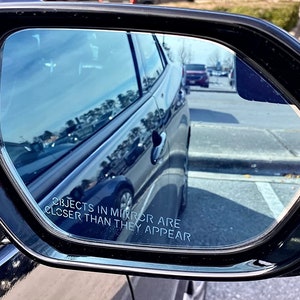 May include: A black car side view mirror with a blue tinted glass. The mirror reflects a car parked in the background. The mirror has a warning label that reads "Objects in mirror are closer than they appear."