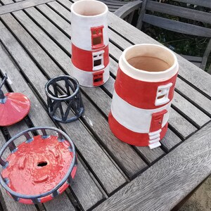 K&ouml;nnte beinhalten: Ein Set aus vier Keramik-Pflanzgef&auml;&szlig;en in Leuchturmform in Rot und Wei&szlig; mit schwarzen Akzenten. Die Pflanzgef&auml;&szlig;e sind in verschiedenen Gr&ouml;&szlig;en und Formen, darunter ein hoher Turm, ein niedrigerer Turm, eine runde Basis und eine kleine kuppelf&ouml;rmige Spitze.