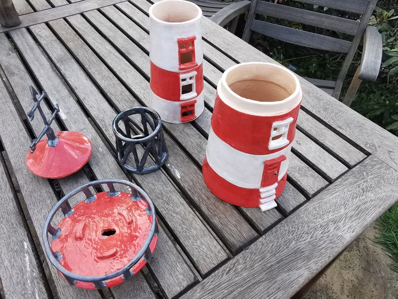 K&ouml;nnte beinhalten: Ein Set aus vier Keramik-Pflanzgef&auml;&szlig;en in Leuchturmform in Rot und Wei&szlig; mit schwarzen Akzenten. Die Pflanzgef&auml;&szlig;e sind in verschiedenen Gr&ouml;&szlig;en und Formen, darunter ein hoher Turm, ein niedrigerer Turm, eine runde Basis und eine kleine kuppelf&ouml;rmige Spitze.
