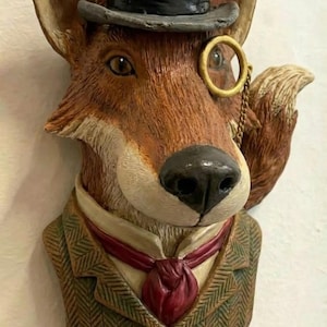 May include: A whimsical fox sculpture with a top hat, monocle, and tweed jacket. The fox has a brown and white face, a black nose, and is wearing a dark gray top hat. The jacket is green with a red tie. The sculpture is a unique decorative item.