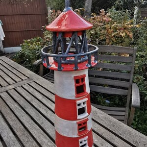 K&ouml;nnte beinhalten: Ein roter und wei&szlig;er Keramik-Leuchtturm mit einem schwarzen Metalldach und einer Wetterfahne. Der Leuchtturm hat eine kleine T&uuml;r und Fenster.