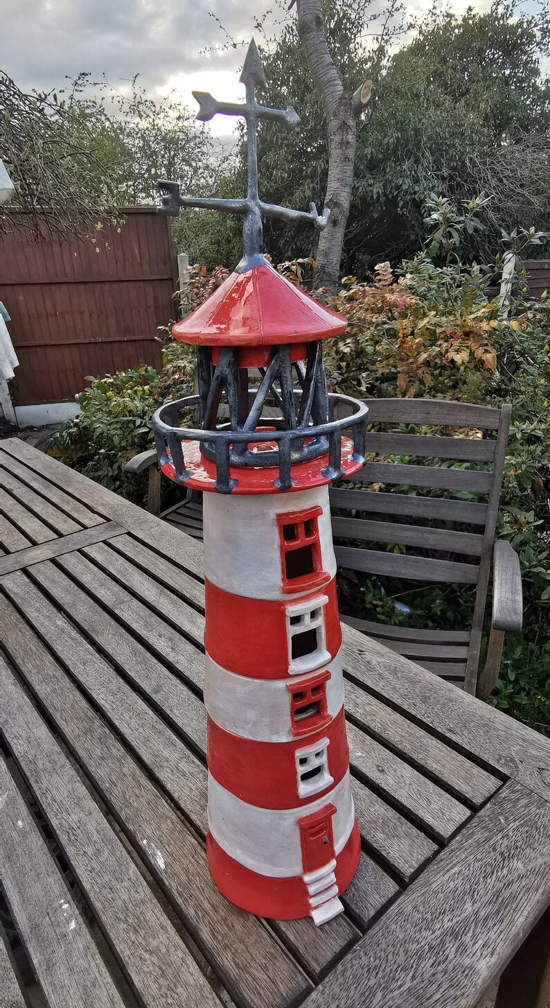 K&ouml;nnte beinhalten: Ein roter und wei&szlig;er Keramik-Leuchtturm mit einem schwarzen Metalldach und einer Wetterfahne. Der Leuchtturm hat eine kleine T&uuml;r und Fenster.