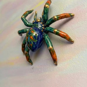 May include: A ceramic spider figurine. It has a blue body, green legs, and orange and gold accents. The spider's body has a swirling pattern of orange and green. The legs are green with orange tips and gold details. The background is a soft, pastel colour.