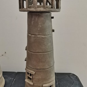 K&ouml;nnte beinhalten: Eine handgefertigte Keramik-Leuchtturm-Figur mit detailliertem Design. Der Leuchtturm ist in hellbraun bemalt und hat ein kleines Fenster und eine T&uuml;r. Der Leuchtturm steht auf einer schwarzen Oberfl&auml;che.
