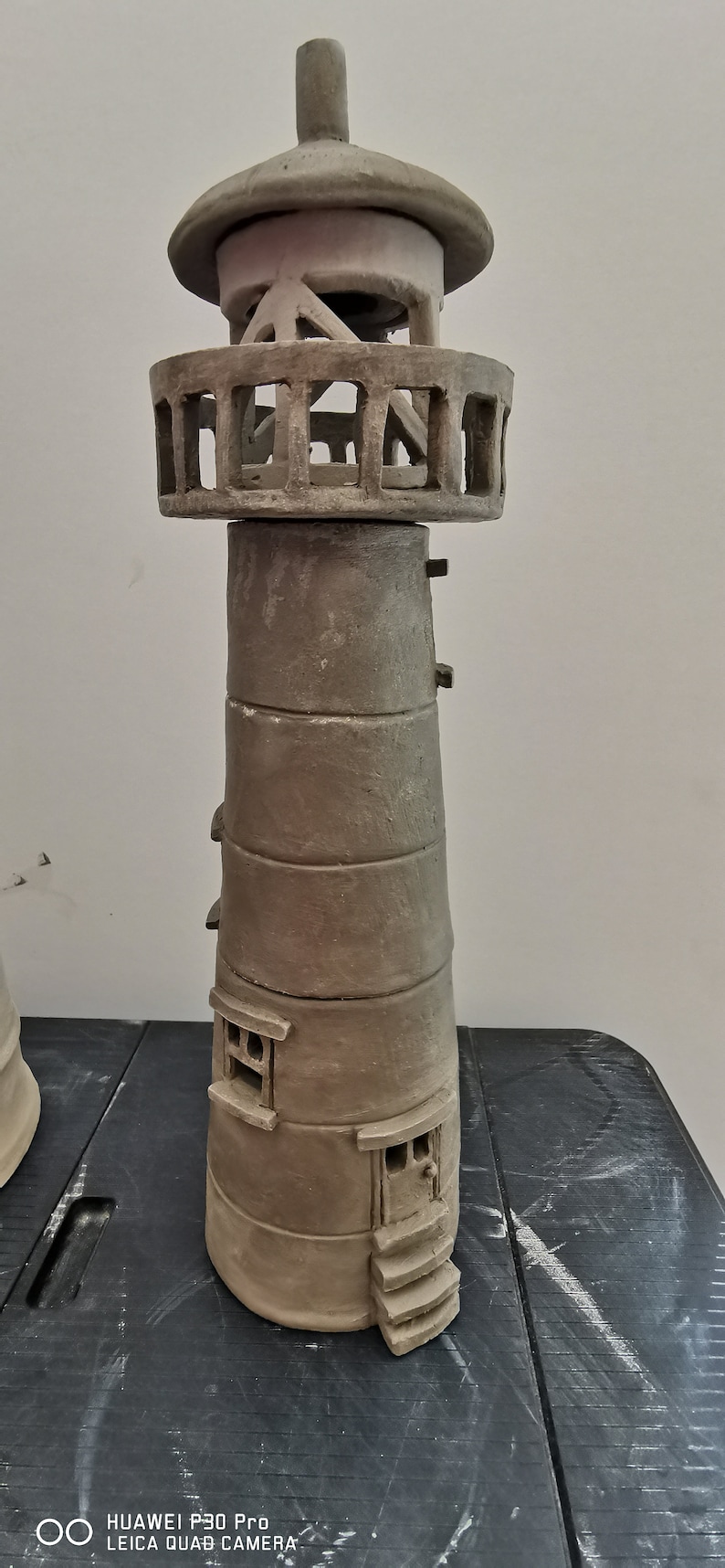 K&ouml;nnte beinhalten: Eine handgefertigte Keramik-Leuchtturm-Figur mit detailliertem Design. Der Leuchtturm ist in hellbraun bemalt und hat ein kleines Fenster und eine T&uuml;r. Der Leuchtturm steht auf einer schwarzen Oberfl&auml;che.