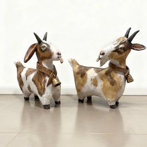 Puede incluir: Dos figuras decorativas de cabras. Las cabras tienen cuerpos manchados marrones y blancos, cuernos marrones y una campana alrededor del cuello. Una cabra saca la lengua. Las figuras están sobre una superficie clara.