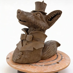 May include: A brown clay sculpture of a fox wearing a top hat, coat, and bow tie. The fox is posed on a wooden wheel. The sculpture has detailed textures and features, including a feather on the hat and buttons on the coat.