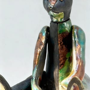 Puede incluir: Escultura de oso de peluche de cerámica con un acabado brillante e iridiscente. El oso presenta una mezcla de colores, incluyendo negro, verde y bronce. El oso está sentado con los brazos y las piernas extendidos.