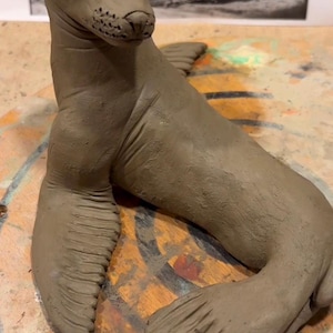 May include: A handmade clay sculpture of a seal in a relaxed pose. The seal is a light brown colour, with detailed flippers and a textured body. The sculpture is set against a blurred background with a black and white image of a seal.