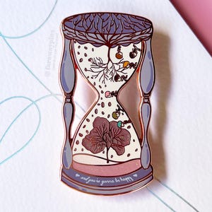 Puede incluir: Un pin de esmalte de reloj de arena de oro rosa con un reloj de arena morado y blanco. El reloj de arena está lleno de un árbol y flores. El texto "and you're gonna be happy" está en la parte inferior del reloj de arena.
