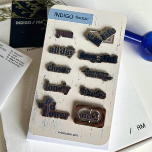 May include: A set of 10 silver and blue enamel pins with different words and designs. The pins are arranged on a white card with the words "INDIGO TRACKLIST" and "interactive pins" printed on it.