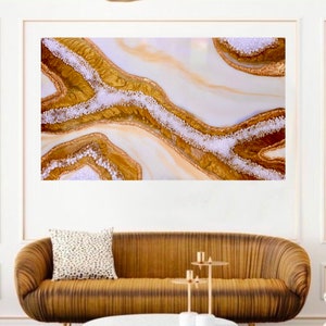 May include: Abstract art print featuring a white background with gold and white swirls and glitter. The print is displayed on a wall above a brown couch and a white coffee table.