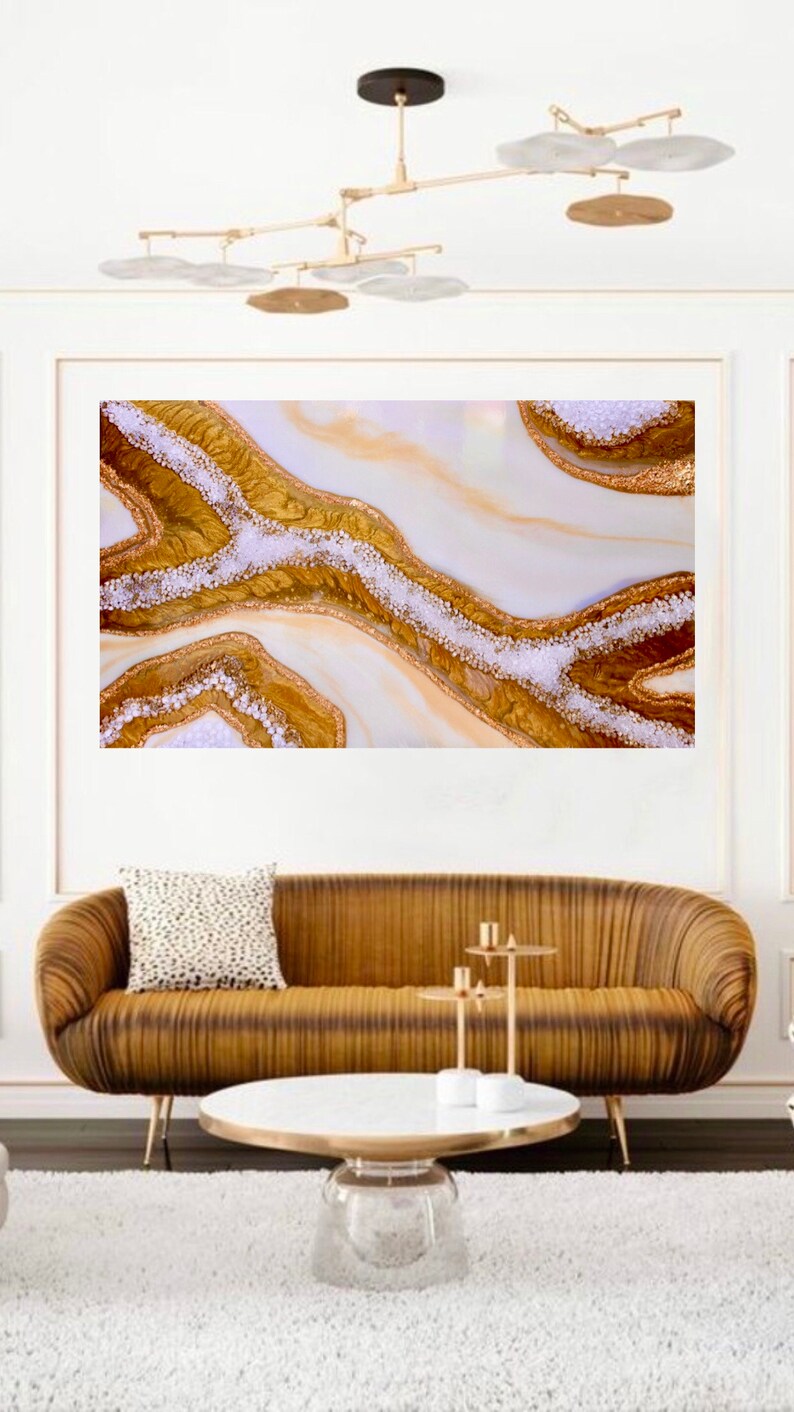 May include: Abstract art print featuring a white background with gold and white swirls and glitter. The print is displayed on a wall above a brown couch and a white coffee table.