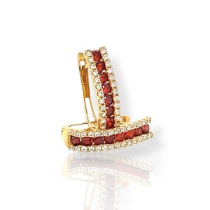 May include: A pair of gold-colored hoop earrings. Each earring features a row of small, red gemstones and a row of clear, white gemstones. The earrings are displayed on a white surface with a reflection.