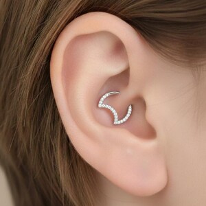 May include: A silver-toned ear piercing featuring a curved design, embellished with small, sparkling clear stones. The jewelry is set within the ear's cartilage, showcasing a delicate and elegant aesthetic.
