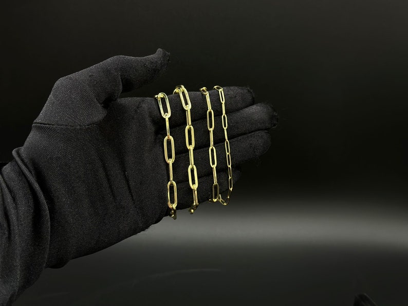 May include: Four gold chain necklaces with a link style that resembles a paperclip. The chains are arranged in a stack, with the largest chain at the bottom and the smallest chain at the top.