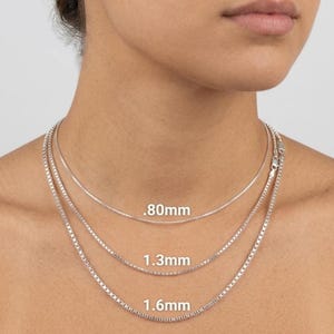 May include: Three silver box chain necklaces of varying thicknesses are displayed. The thinnest chain is .80mm, the middle chain is 1.3mm, and the thickest chain is 1.6mm. The necklaces are around a person's neck.