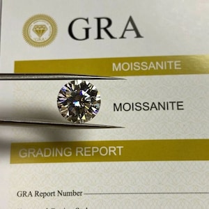 May include: A close-up of a round, clear, colorless gemstone with a brilliant cut. The gemstone is held in tweezers and is labeled "Moissanite" on a white card with gold lettering. The card also has a logo with the letters "GRA" in a circle.