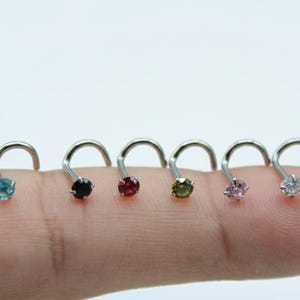 Puede incluir: Seis piercings nasales con postes curvos, cada uno con una gema de diferente color. Las gemas incluyen azul, negro, rojo, verde, rosa y transparente. Las joyas se muestran en un dedo sobre un fondo blanco.