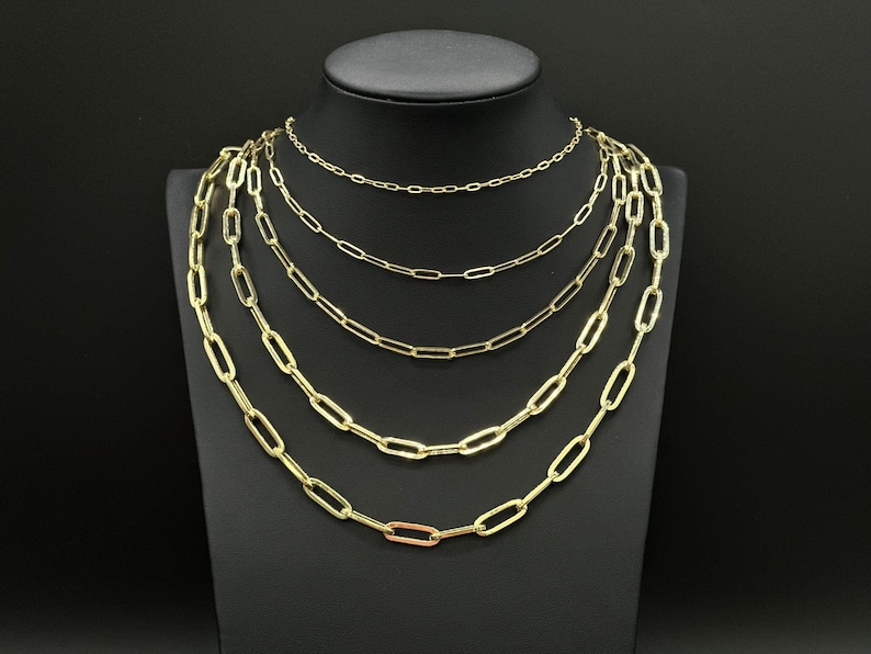May include: A set of five gold chain necklaces with different lengths, all featuring a classic oval link design. The chains are layered on a black mannequin bust.