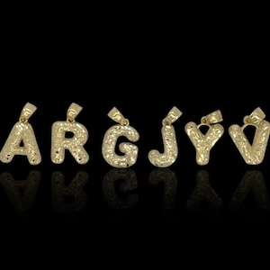 May include: Gold-colored letter pendants spelling out "ÁRGJV" against a black background. Each pendant has a textured surface and a loop for a chain. The letters are in a bubble font.