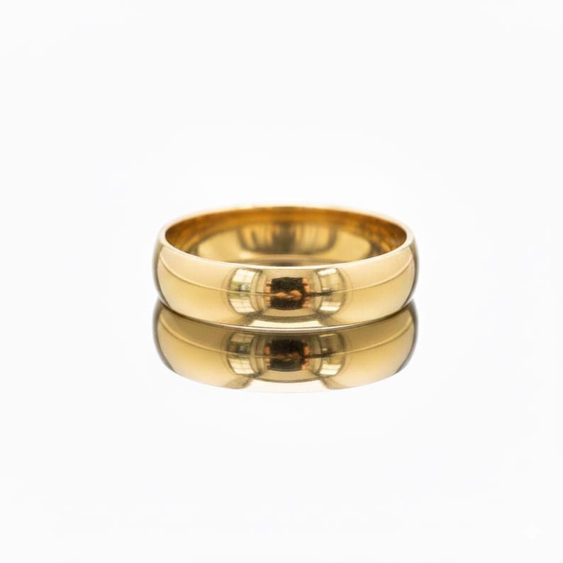 May include: A gold band ring with a polished finish. The ring is round and reflects the light, creating a bright, shiny appearance. The ring is sitting on a reflective surface, creating a mirrored image.